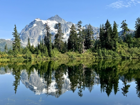 Scenic photo of A mountain with some patches of snow and trees along a waterbody. The water is still and reflects the mountain, sky and trees.