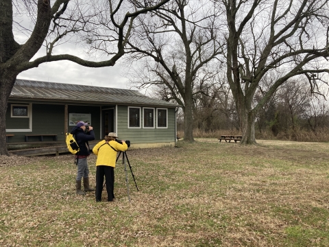 Two people stand outside near trees and a building looking through binoculars and a scope