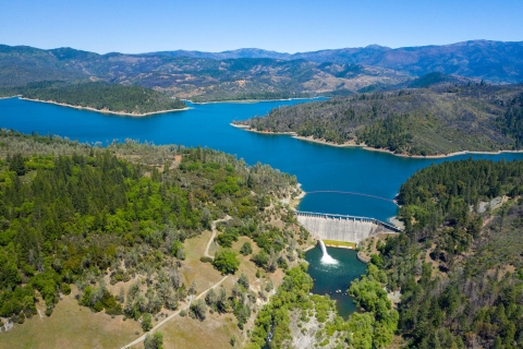 Aerial view of a large bright blue lake surrounded by forested mountains. A large dam spans the front section of the lake.