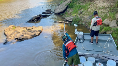 Two men standing in a boat in a river. One man is measuring the depth of the water.