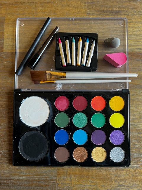 a tray with 18 shallow pots of face paint in varied colors, 6 face paint crayons in varied colors, two paint brushes, two makeup sponges, and two liquid eyeliner pens in black and brown.