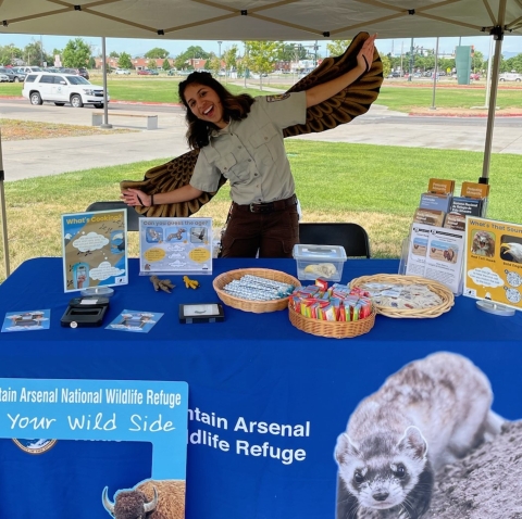 A person smiling and wearing brown wings while standing behind a blue table full of trinkets and educational materials.