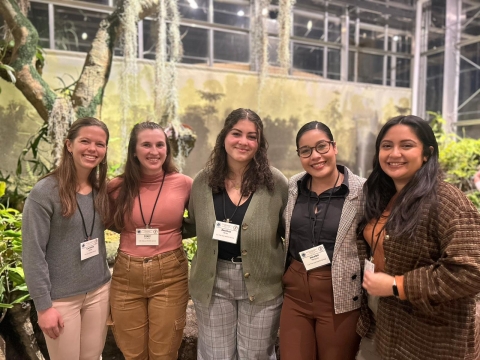 5 young people stand smiling together in front of indoor foliage. They all wear white lanyards with their names displayed