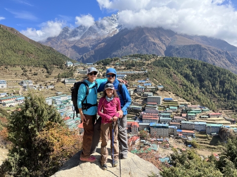 Two adults and a child pose in front of a colorful marketplace in with big mountains, clouds, and blue sky in the background. 