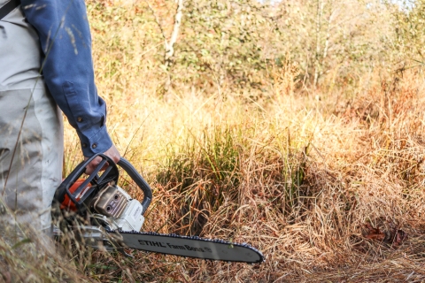 Person standing among tall grass holding a chainsaw. Trees in the distance,