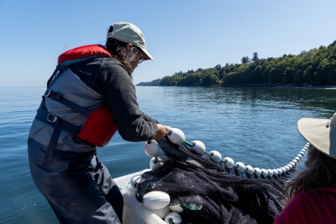 A service intern in waders and a PDF pulls a net out of the water onto a boat. The harbor and forested shoreline are seen in the background.