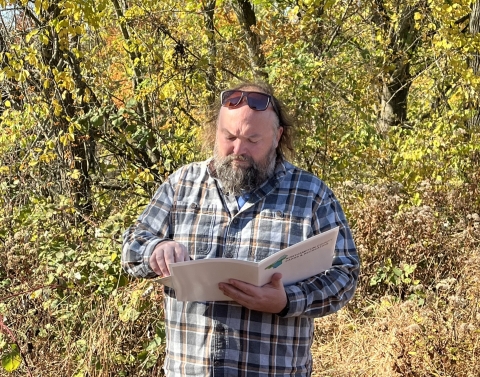 a white man with a brown-and-gray mustache and beard, wearing brown sunglasses on his head and a gray-and-blue plaid shirt holds a white folder and reads from it, surrounded by golden shrubs and trees