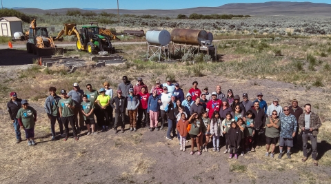 A group of tribe members take a photo from a low flying drone. There are tractors, oil tanks, and sagebrush and mountains in the background.