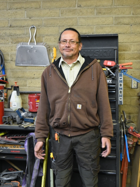 A man wearing a brown sweatshirt and glasses stands in a maintenance shop