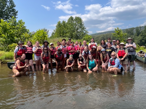 Many smiling people wearing life jackets standing in shallow water with canoes and a forested horizon.