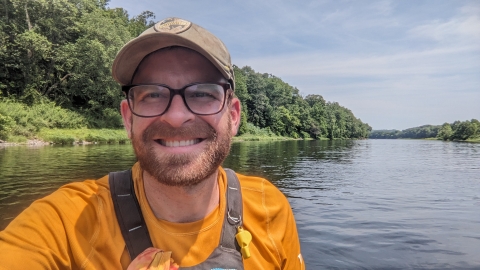 a smiling white man with large brown-rimmed glasses and a light brown mustache and beard, wearing a tan ball cap and a bright gold shirt with water and forest behind him