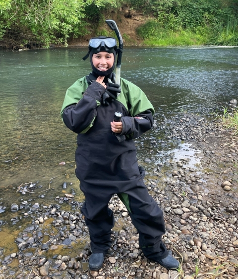 Service intern stands by the side of a river wearing a drysuit and snorkel gear. Rocky river shore, river, and tree foliage seen in the background.