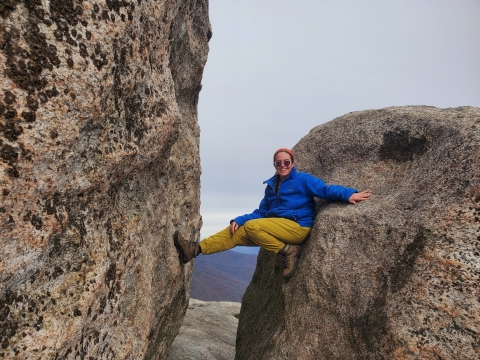 A young woman in a blue jacket smiles as she rests wedged between two boulders. The sky is grey and mountains are visible in the background.