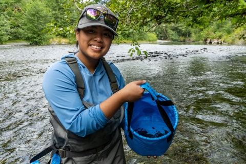 A Pathways employee holds up a bucket full of small Sculpin. A river and tree foliage can be seen in the background. 