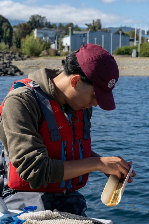 A service intern wearing a PFD measures a fish against a ruler. Water and rocky beach shore can be seen in the background. 