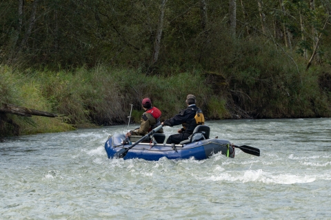 A service intern and USFWS employee float down a river in a two-man inflatable raft. They are wearing dry suits and PFDs, trees and ground foliage can be seen in the background.