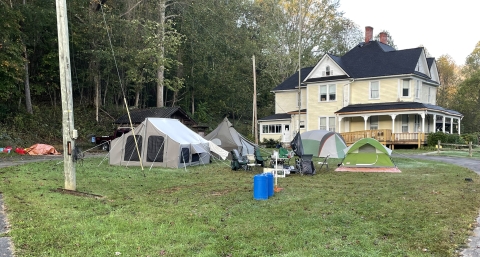 Looking across a green lawn with trees in the background is a large yellow house with a wrap around porch. In the foreground, near the house are several tents set up providing a place for storm volunteers to stay. 