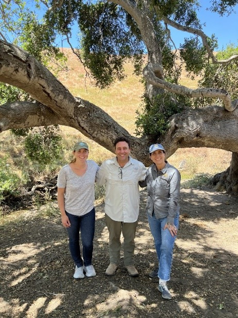 Three people stand in front of a large tree. The center person is a man with a white shirt and green pants and the two women on the end wear USFWS caps, gray shirts, jeans and sneakers. 