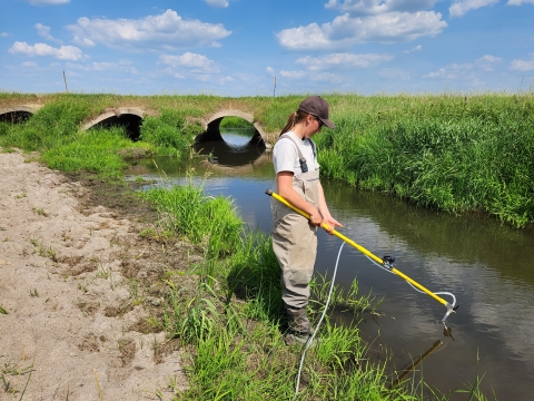 Woman in waders holds a pole and sticks it in the water. It is a summers day.