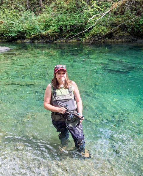 A service member wearing waders stands in a river holding a camera.