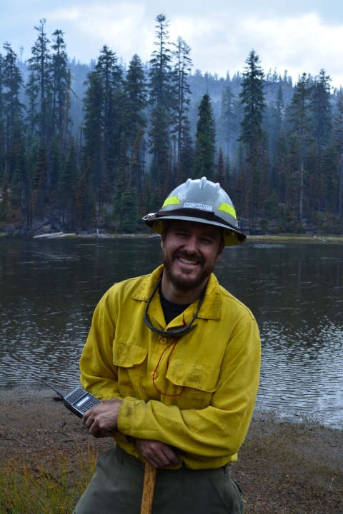 A firefighter dressed in yellow protective clothing and hard hat, smiles and leans on their tool with a radio in hand. Behind him is a mountain lake lined with tall evergreen trees. The sky is hazy with smoke.