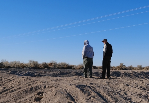 Two men stand facing away from the camera and looking left. They stand over newly excavated dirt ponds..