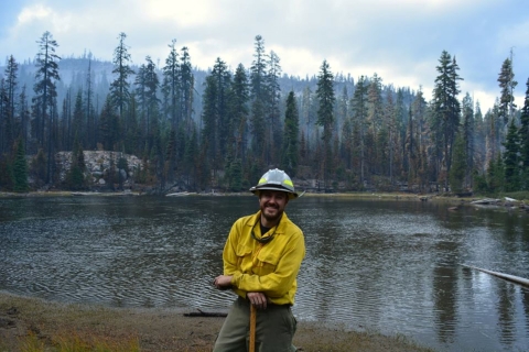 A firefighter dressed in Nomex protective clothing and hard hat, smiles and leans on their tool. Behind them is a lake and tall evergreen trees. The sky is hazy with smoke.