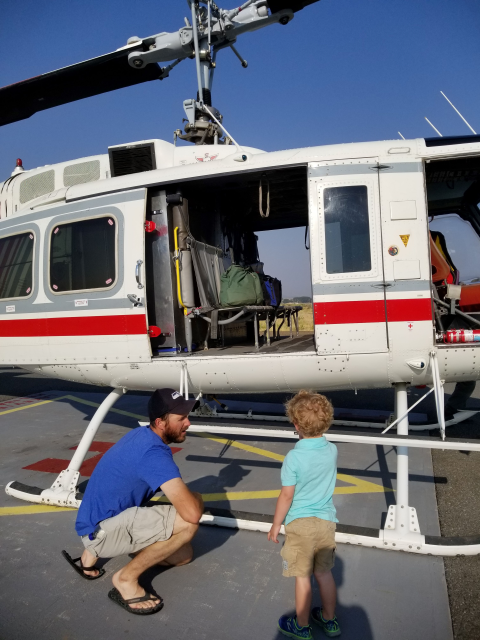 A dad crouches down to his young son as he shows him a firefighting helicopter on the tarmac. The helicopter is white with a red stripe, and has it's doors open.