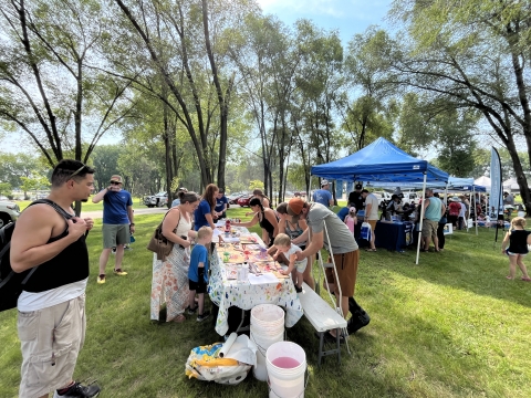 Children paint at a table while parents assist them outdoors. There are multiple canopies set up and crowds of people talk to vendors in the distance.