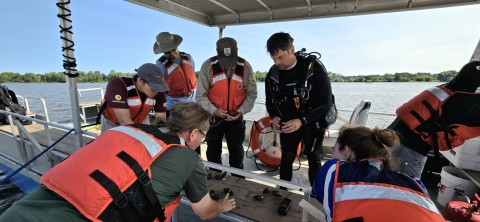 A group of people in life vests and a diver lean in to look at several mussels on the deck of a boat.