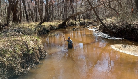 Man in waders walks in murky water surveying water depth