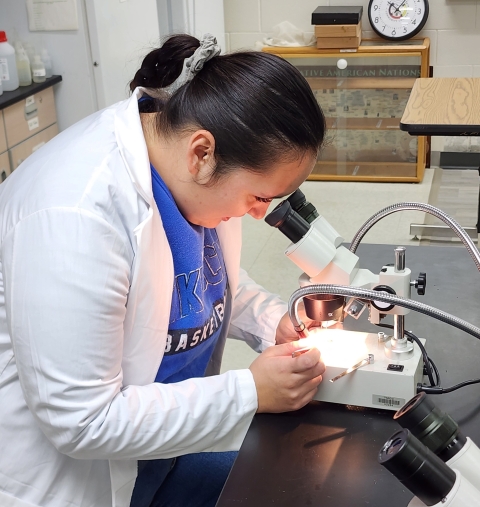 student in a white coat looking into a microscope in the lab