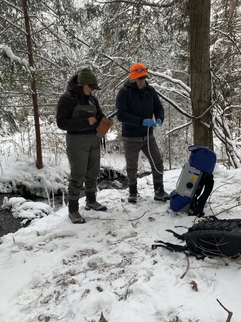 Image of 2 people in winter attire standing next to a tree and blue backpack in the snow.