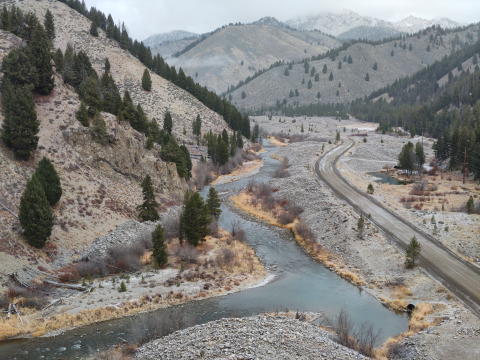 An aerial view over a winding river with a road along one side and mountains with evergreen trees on the other. there is a light dusting of snow on the road and terrain.