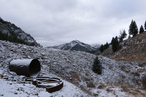 Old remnants of rusty equipment lay in a pile of rocks and rubble dusted with snow. Mountains and trees are in the background.