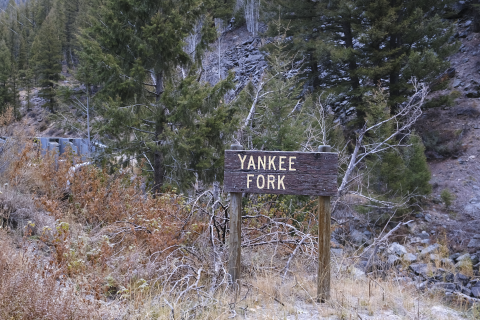 A wooden sign says "Yankee Fork" with pine trees and mountains behind it