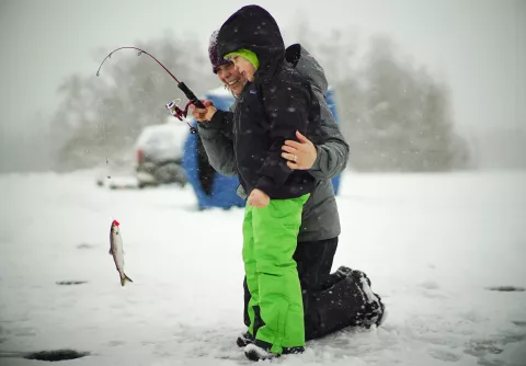 A woman and young boy stand on a frozen lake. The woman is helping the boy hold up a fish they have just caught from a hole in the ice.