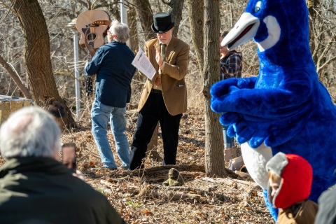 Two men, one holding a guitar, sing next to a blue goose mascot.