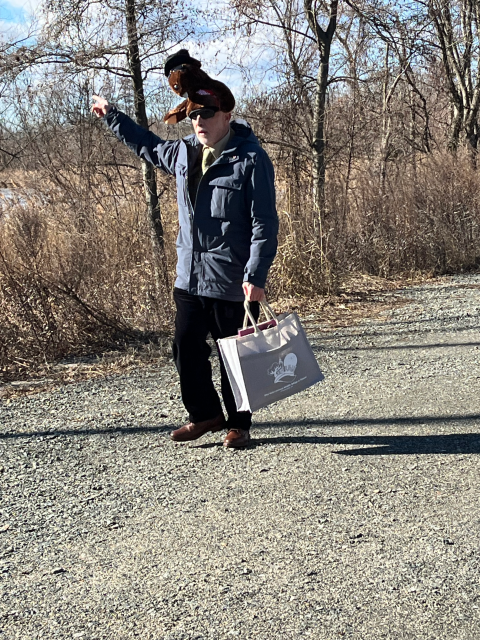 Man wearing stuffed groundhog hat stands outdoors on a gravelly road and points at a tree.