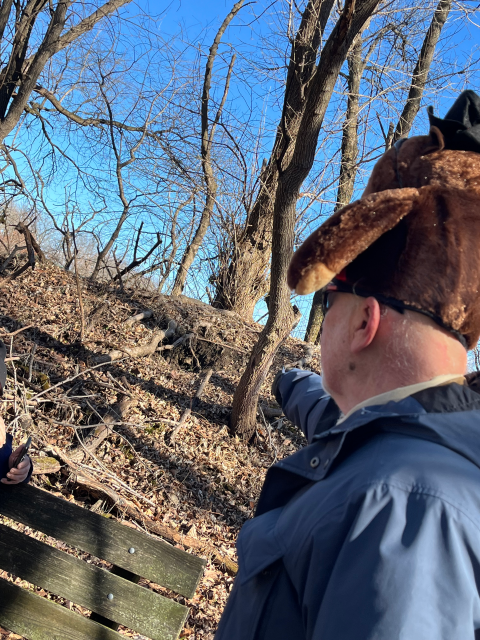 Man wearing a stuffed groundhog hat points at a groundhog burrow.