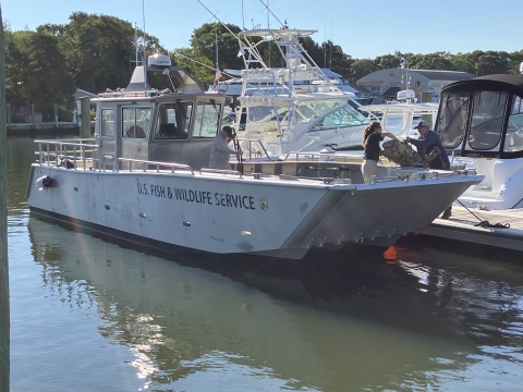 A person standing on a dock hands a large bag to a person standing on the deck of a metal boat