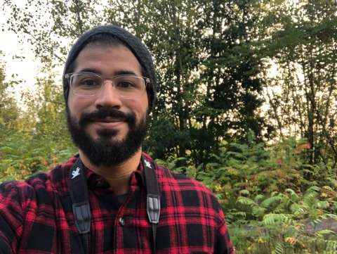 a bearded man with glasses and a beanie hat smiles while standing in a lush green forest. A black binocular strap hangs around his shoulders