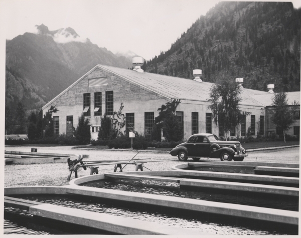 Black and white photo of oval shaped fish ponds, car, hatchery building, and mountains