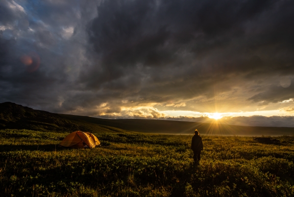 A person looks on with a yellow tent in the foreground and a sun setting behind a hill under a cloudy sky in the background