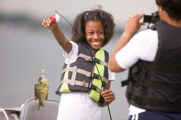 A young girl holds a fish that she caught on the end of her fishing line as her mother takes a photograph of her. 