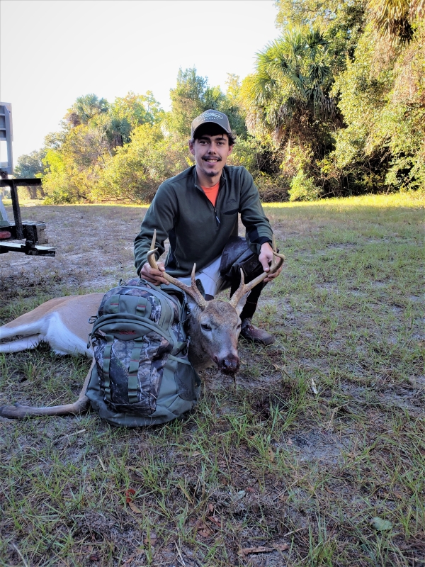 Young archer kneels on the ground with the six-point buck he harvested on Bulls Island. The morning sun shines brightly on the trees behind the hunter 