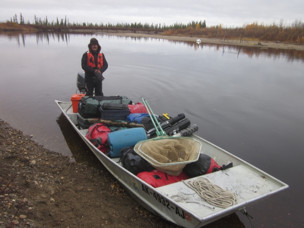 a man stands in the stern of a small open motorboat which is loaded with outdoor gear