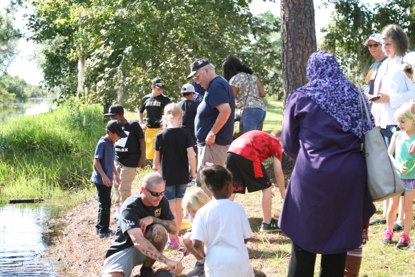 AS crowd of people, children and adults, on the edge of the water. Some people crouched down looking at something.