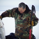 A Native Alaskan wearing a warm camo jacket pulling a white and silver fish out of the ice