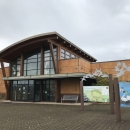 Large building with glass windows and tan siding; angled roof; sign that says "visitor center"; interpretive panels near building; stone paved ground